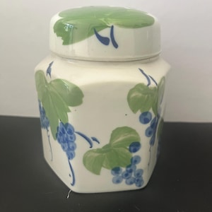 May include: A white ceramic jar with a green and blue grape design. The jar has a lid with a green top and a white base.