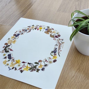 Op de afbeelding: Een witte kaart met een krans van gedroogde bloemen en bladeren in tinten bruin, geel, paars en rood. De krans is in een cirkel op de kaart gerangschikt.