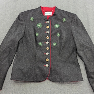 Puede incluir: Una chaqueta de lana gris oscuro con forro rojo y flores bordadas verdes. La chaqueta tiene un cuello alto y cierre de botones.