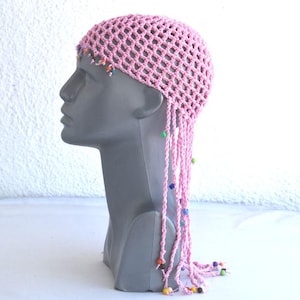 Puede incluir: Sombrero de crochet rosa con flecos de cuentas. El sombrero está hecho de un tejido fino y abierto y tiene un ajuste holgado. El fleco está hecho de cuentas de colores y cuelga de la parte posterior del sombrero.