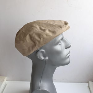 May include: A beige flat cap made from a soft material. The cap is displayed on a grey mannequin head, highlighting its shape and design. It features a rounded crown and a small, rigid peak.