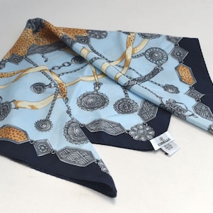 May include: A blue and navy blue silk scarf with a pattern of silver chains and medallions. The scarf has a label that reads "Coccinella".