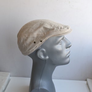 May include: A beige cotton newsboy cap with a slightly curved brim and two brass eyelets on the side.