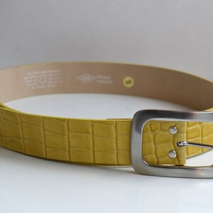 Op de afbeelding: Mosterdgele riem met een textuur die op krokodillenleer lijkt. De riem heeft een zilverkleurige, rechthoekige gesp met een enkele pin. De binnenkant van de riem is lichtbruin.