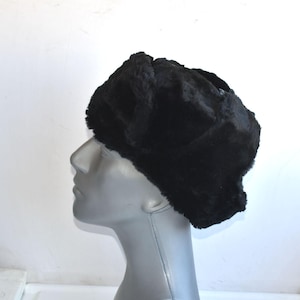 May include: A black faux fur hat with a rounded crown and ear flaps. The hat is displayed on a mannequin head against a white backdrop. The hat's fur looks soft and plush.