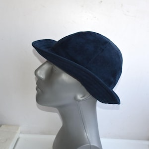 May include: A dark navy blue bucket hat with a rounded crown and a short, downward-sloping brim. The hat features a smooth, uniform texture and is displayed on a neutral-toned mannequin head to showcase its shape and fit from a side profile.