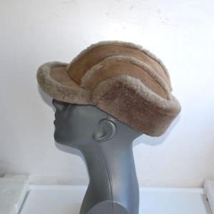 May include: A tan-colored hat with a curved brim and ear flaps. The hat is made of a soft, fuzzy material, likely shearling, and is designed to provide warmth. The hat is displayed on a mannequin head.