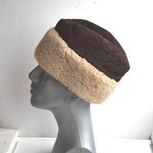 Op de afbeelding: Een bruine wintermuts met een beige, pluizige rand. De hoed is gemaakt van een donkerbruin, getextureerd materiaal aan de bovenkant. De hoed wordt getoond op een mannequinhoofd.