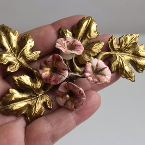 以下が含まれることがあります： ピンクと白の磁器の花がついた金色の金属製のフラワーモチーフ。花は朝顔の形をしていて、金色のアクセントが施されています。葉は細かくリアルです。