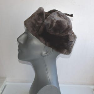 May include: A brown faux fur hat with ear flaps, displayed on a mannequin head. The hat has a soft, textured appearance and a dark brown colour. The ear flaps are folded up. The hat is designed to provide warmth and protection from the cold.