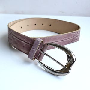 May include: A mauve belt with a textured crocodile-embossed pattern and a smooth, light-colored interior lining. The belt features a polished silver-tone metal buckle with a rounded, contoured frame and a single matching belt loop.
