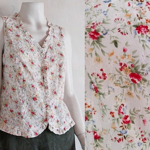Könnte beinhalten: Eine weiße, ärmellose Weste mit einem Blumendruck mit roten, rosa, gelben und blauen Blumen. Die Weste hat einen Rüschenkragen und einen Knopfverschluss.