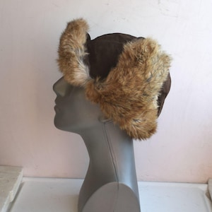 May include: A brown suede and fur trapper hat with ear flaps. The hat has a brown suede exterior and a fur lining. The ear flaps are made of fur and can be tied at the top of the head.