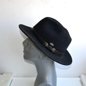 Könnte beinhalten: Ein schwarzer Fedora-Hut mit einem dunkelbraunen Lederband um die Krone. Das Band ist mit zwei dekorativen Schlaufen in bronzefarbener Metalloptik und einem kleinen ovalen, goldfarbenen Metallemblem an der Seite des Hutes verziert.