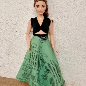 May include: A doll wearing a black crop top with a knot detail and a green skirt with a black print. The skirt has a wrap style and is long and flowing. The doll is wearing black shoes and has brown hair.