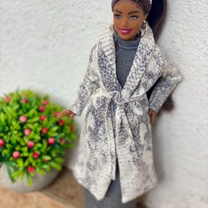 May include: A fashion doll wearing a gray dress and a long coat with a white and gray pattern. The doll has sunglasses on her head and hoop earrings. A green plant with red berries is in the background.
