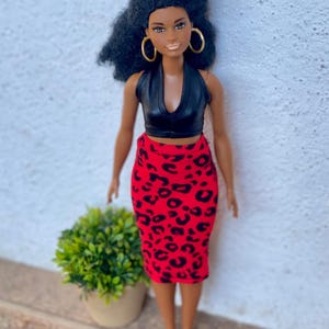 May include: A fashion doll with dark skin, wearing a black halter top and a red pencil skirt with a black leopard print. The doll has black curly hair, gold hoop earrings, and black heels. A small green plant in a beige pot is in the background.