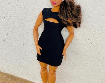 Puede incluir: Una muñeca de moda con un vestido negro sin mangas con un detalle de corte. La muñeca tiene el pelo castaño, los ojos marrones y lleva tacones altos negros.
