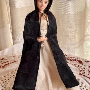 May include: A doll with brown hair wearing a long, cream-colored dress with a lace-trimmed bodice and a black hooded cloak. The cloak features a subtle, dark-on-dark floral pattern and is secured at the neck with a thin, braided brown cord.