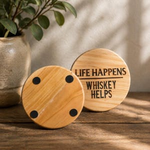 Puede incluir: Dos posavasos redondos de madera clara con el texto negro "LIFE HAPPENS WHISKEY HELPS". Uno boca arriba, el otro boca abajo, mostrando cuatro almohadillas negras. Sobre una superficie de madera.