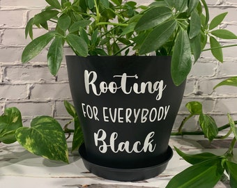 Peut inclure: Un pot de plante noir avec du texte blanc qui dit "Rooting for Everybody Black". Le pot est rempli d'une plante verte feuillue.