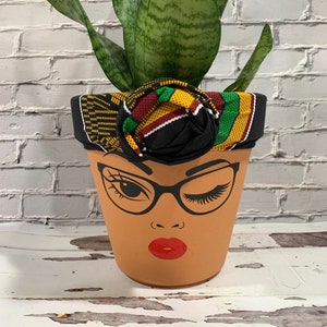 May include: A terracotta flower pot with a painted face design, featuring glasses, a winking eye, and red lips. The pot is adorned with a black fabric band and a colorful patterned fabric flower, and it holds a green plant with long, pointed leaves.