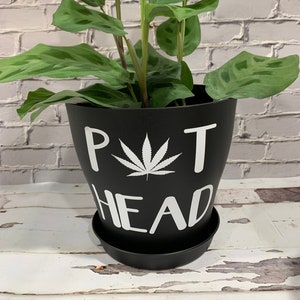 May include: Black plant pot with a white marijuana leaf and the text "Pot Head" on the side. The pot is sitting on a white wooden surface.