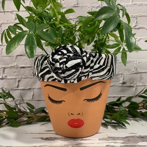May include: A terracotta planter with a black and white zebra print fabric wrapped around the top. The planter is painted with a brown face, closed eyes, and red lips.
