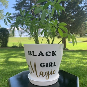 Peut inclure: Pot de plante blanc avec le texte "BLACK GIRL MAGIC" en lettres noires et dor&eacute;es. Le pot est pos&eacute; sur une table hexagonale noire.