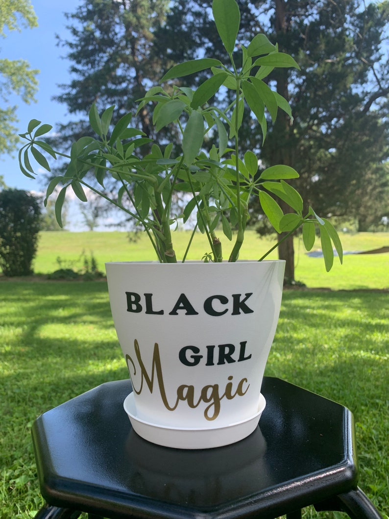 Peut inclure: Pot de plante blanc avec le texte "BLACK GIRL MAGIC" en lettres noires et dor&eacute;es. Le pot est pos&eacute; sur une table hexagonale noire.
