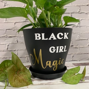 Peut inclure: Un pot de plante noir avec le texte "BLACK GIRL Magic" en lettres blanches et dor&eacute;es. Une plante verte pousse dans le pot.