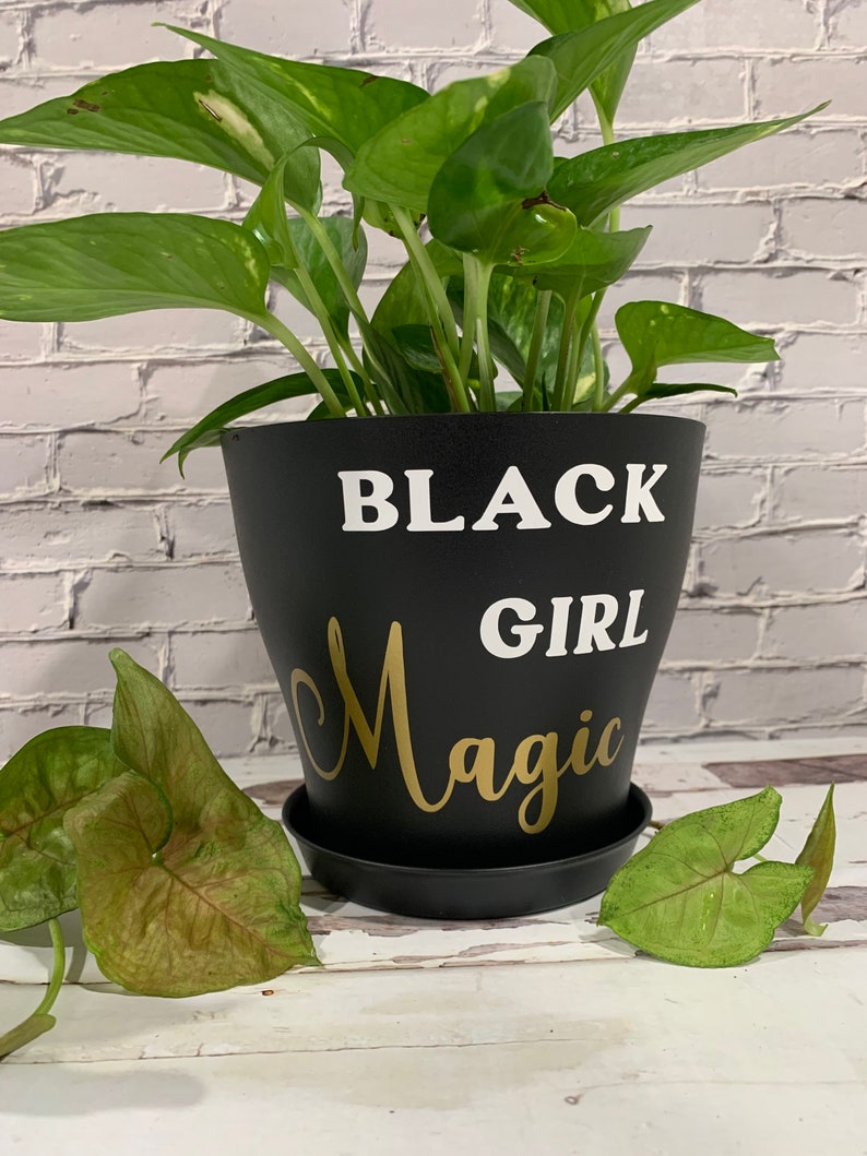 Peut inclure: Un pot de plante noir avec le texte "BLACK GIRL Magic" en lettres blanches et dor&eacute;es. Une plante verte pousse dans le pot.