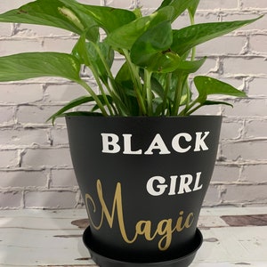 Peut inclure: Un pot de plante noir avec les mots "Black Girl Magic" &eacute;crits en blanc et or. Une plante verte en pot est dans le pot.