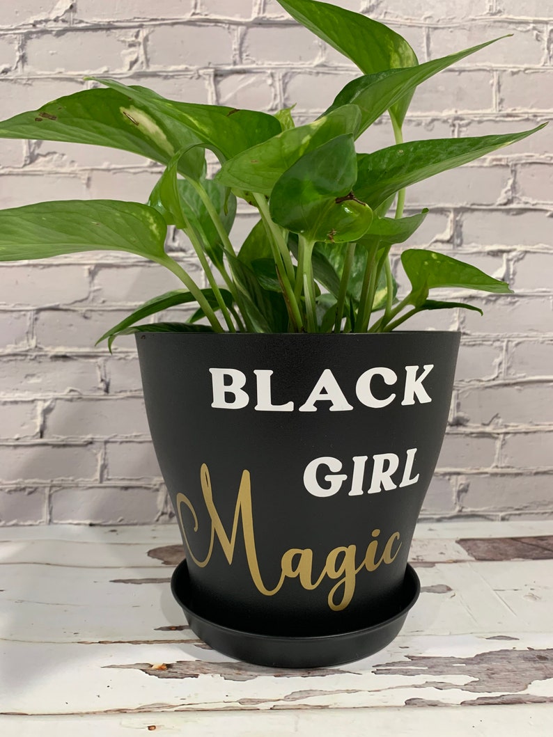 Peut inclure: Un pot de plante noir avec les mots "Black Girl Magic" &eacute;crits en blanc et or. Une plante verte en pot est dans le pot.
