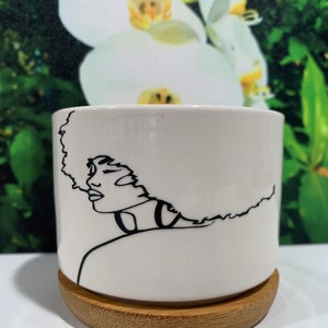 May include: White ceramic planter with a black line drawing of a woman with an afro. The planter has a wooden base.