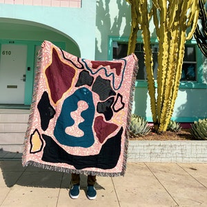May include: A woven tapestry throw blanket with a pink, black, teal, and brown abstract design. The blanket is being held up in front of a building with a cactus in the background.
