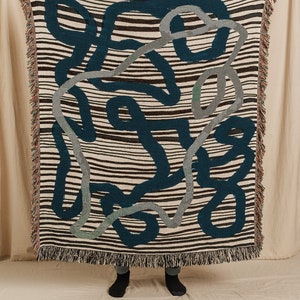 Puede incluir: Una manta tejida con un fondo a rayas y un patr&oacute;n de formas abstractas en azul verdoso, gris y marr&oacute;n.