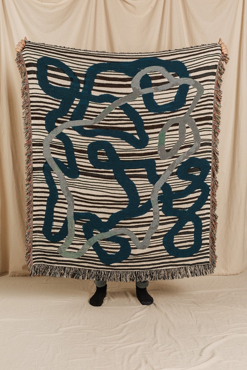 Puede incluir: Una manta tejida con un fondo a rayas y un patr&oacute;n de formas abstractas en azul verdoso, gris y marr&oacute;n.