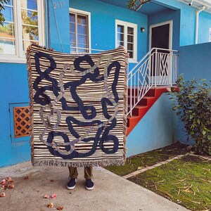 Puede incluir: Una persona sostiene un tapiz tejido con un fondo a rayas blanco y negro y un gran dise&ntilde;o abstracto en azul y verde. El tapiz est&aacute; colgado frente a un edificio azul con una escalera roja.
