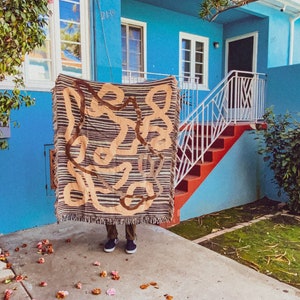 Puede incluir: Una persona de pie frente a una casa azul con una tapicer&iacute;a tejida marr&oacute;n y beige colgada frente a ella. La casa tiene una barandilla blanca y escalones rojos que conducen a la puerta principal. Los n&uacute;meros de la casa 2139 y 2145 son visibles en la casa.