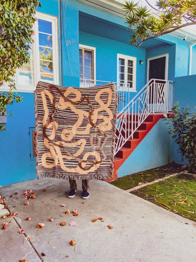 Puede incluir: Una persona de pie frente a una casa azul con una tapicer&iacute;a tejida marr&oacute;n y beige colgada frente a ella. La casa tiene una barandilla blanca y escalones rojos que conducen a la puerta principal. Los n&uacute;meros de la casa 2139 y 2145 son visibles en la casa.