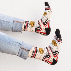 May include: A pair of white socks with a black, red, and yellow abstract pattern. The socks are worn by a person wearing blue jeans.