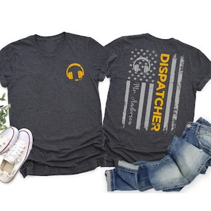 May include: A dark heather grey t-shirt with a yellow headset graphic on the front. The back of the shirt features a distressed American flag design with the text "DISPATCHER" in yellow and "Mr. Anderson" in grey.