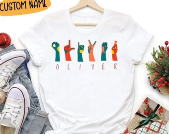 Puede incluir: Una camiseta blanca de manga corta con un gráfico de seis manos haciendo las letras del alfabeto manual para deletrear el nombre OLIVER en texto rojo debajo. Las manos están coloreadas en tonos verde azulado, naranja y rojo con detalles amarillos.