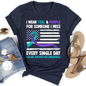 May include: Navy blue t-shirt with the text "I WEAR TEAL & PURPLE FOR SOMEONE I MISS EVERY SINGLE DAY SUICIDE PREVENTION AWARENESS." The design includes a teal and purple ribbon over a white and blue American flag.