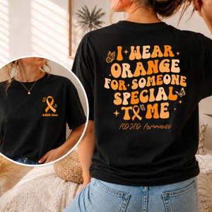 Pode incluir: Uma t-shirt preta com texto e gráficos em laranja. As costas exibem o texto I WEAR ORANGE FOR SOMEONE SPECIAL TO ME com borboletas e uma fita. No peito, vê-se uma pequena fita laranja e o texto ADHD Mom.