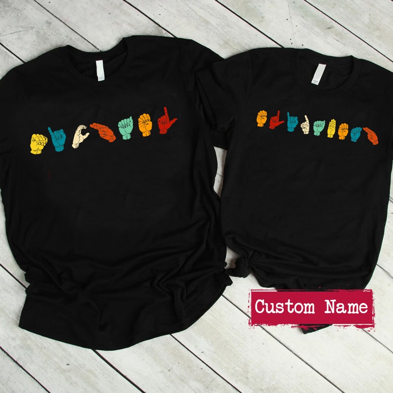 May include: Two black t-shirts with a line of colorful hands signing the word "LOVE" in American Sign Language. The smaller shirt has the text "Custom Name" printed below the design.