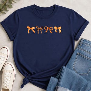 Può includere: Una t-shirt a maniche corte blu navy con una fila orizzontale di quattro fiocchi a nastro arancioni stampati al centro del petto. Il capo presenta uno scollo rotondo e un dettaglio annodato sull'orlo inferiore sul lato sinistro.
