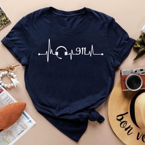 May include: Navy blue t-shirt featuring a white graphic of a heartbeat line with a headset and the number 911. The shirt has rolled-up sleeves and is made of a soft material. The shirt is displayed on a neutral surface.