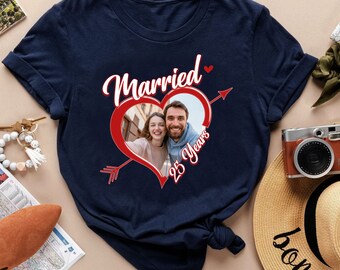 Puede incluir: Una camiseta azul marino con un gráfico de un corazón rojo atravesado por una flecha. Dentro del corazón hay una fotografía de una pareja sonriente. El texto "Married" aparece arriba y "25 Years" al lado del corazón.